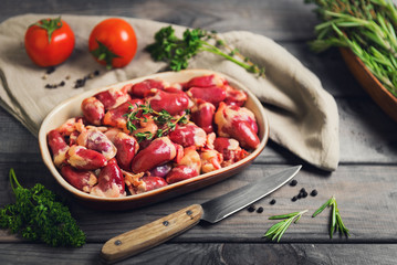 Fresh raw chicken hearts in a ceramic bowl. Seasonings for chicken hearts, thyme, rosemary, parsley, pepper, tomatoes. Rustic wooden gray background.