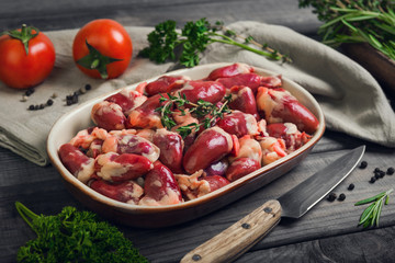 Fresh raw chicken hearts in a ceramic bowl. Seasonings for chicken hearts, thyme, rosemary, parsley, pepper, tomatoes. Rustic wooden gray background.