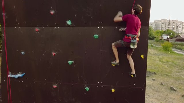 Aerial of climber runs the speed rock climbing track on artificial wall outdoors. Young fast athlete climbs the cliff at competitions. Man on belaying harness performs at bouldering championship.