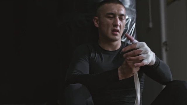 Medium shot of sportsman in compression garments leaning on punching bag and wrapping his hands with elastic bandage before doing boxing workout