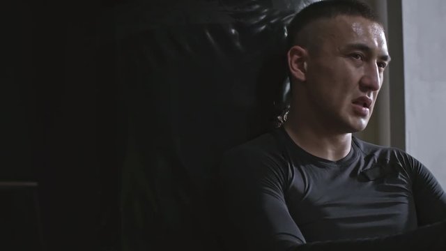 Medium shot of exhausted man relaxing after hard workout at the gym, sitting and leaning on punching bag