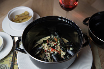 Stewed mussels with vegetables in a saucepan on a served table with dishes and red wine glass