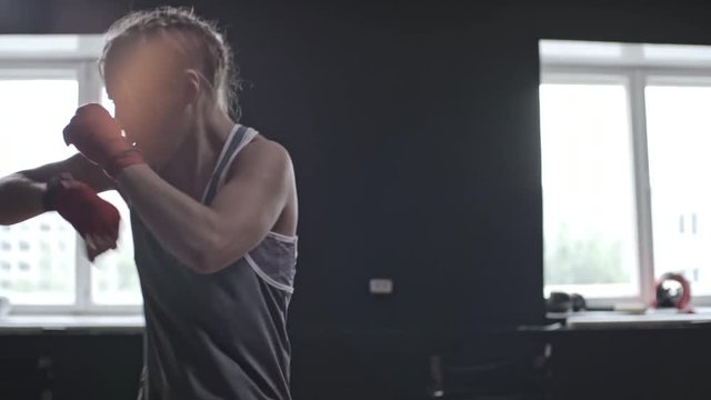 Medium shot of fit young woman with two braids practicing kicks and punches when doing shadow boxing workout in sports studio