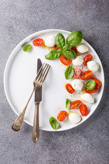 White plate of healthy classic delicious caprese salad with ripe tomatoes and mozzarella cheese with fresh basil leaves on gray concrete background with space for text. Italian food. Top view.