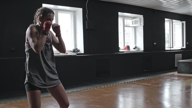 Fit young woman in sports clothes exercising in sports studio and practicing kicks and punches as part of shadow boxing workout
