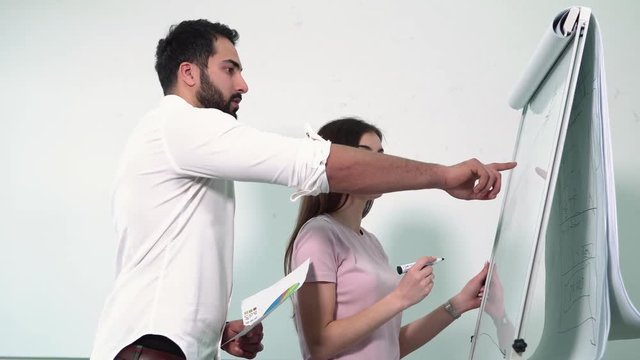 Two apprentices drawing picture on flip chart board, dark-haired man wearing smart white shirt, lovely brunette - pink t-shirt