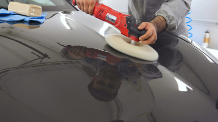 A man in a special suit polishes a gray car body, a tool for polishing cars, into a workshop.