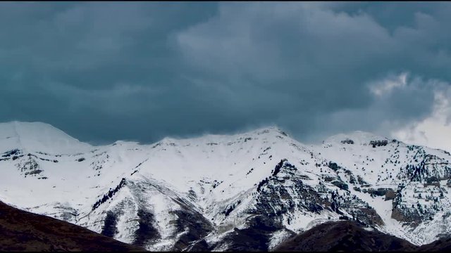 Zooming out from the top of a misty mountain, this time lapse shows a dramatic cloudscape above snow-covered mountains with foothills in the foreground