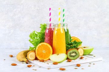 Green, yellow, purple smoothies in currant bottles, parsley, apple, kiwi, orange on a gray table.
