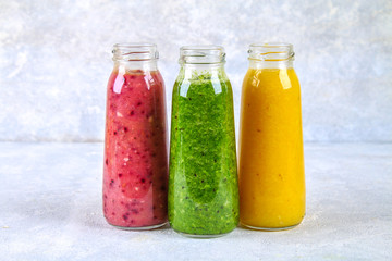 Green, yellow, purple smoothies in bottles of berries, greens, oranges on a white board on a gray table.