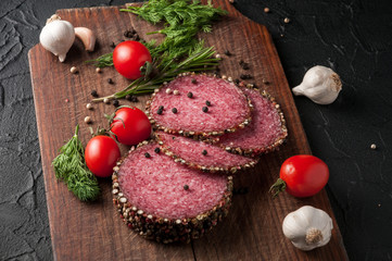 Smoked salami on a black table and a brown board. Sausages with rosemary, garlic and pepper and tomato cherry, dill, parsley.