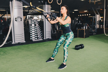 TRX concept. Beautiful lady exercising her muscles with help of suspension trainer sling or suspension straps in gym