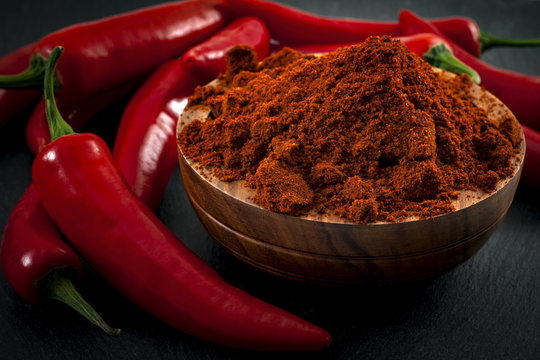 Mexican cuisine and spicy food concept with close up on fine ground red pepper powder in a wooden bowl surrounded by a bunch of hot chilli peppers on a dark background with dramatic light