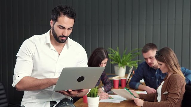 Handsome asian man working with pleasure on silver laptop, standing joyfully before group of office workers inventing brand improvement ideas