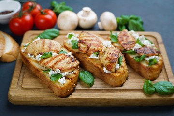 Tasty crunchy italian snack bruschetta with grilled chicken, garnished with ricotta and basil on wooden board