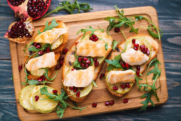 Tasty homemade Italian antipasti bruschetta with grilled chicken and zucchini, garnished with pomegranate seeds and arugula, healthy and delicious snack flat lay on wooden table