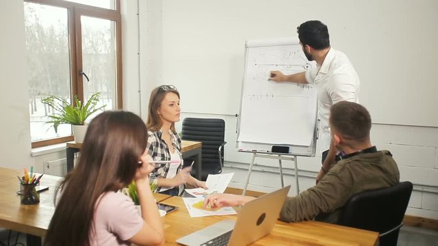 Team of marketing professionals discussing a piece of graph drawn on the flipchart, busy atmosphere in light modern office