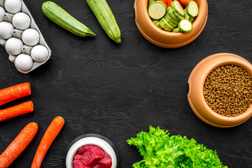 pet care with dry food, vegetables and raw meat for dog or cat in plastic bowl on black background top view space for text