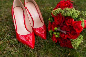  Two golden wedding rings lie on red fashion female shoes on green grass. Wedding details