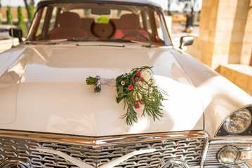 Lush bridal bouquet with white and pink flowers with a lot of greenery lying on the hood of white car of bride and groom.