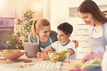 Nice picture. Delighted kid holding tablet in both hands and putting elbows on the table while sitting in the kitchen near mother