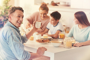 Relax at home. Positive delighted male person keeping smile on his face and holding white cup in left hand while sitting in semi position