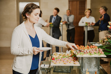 Young businesswoman serving herself with canapes in buffet of restaurant. Business people eating...