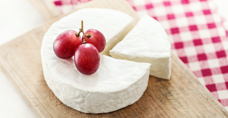 Cheese wheel with a piece of cheese on a wooden board with fruits and red checkered tablecloth. Camembert cheese close up..