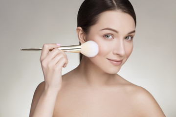 Beautiful brunette caucasian young woman prepare herself, applying powder on her cheeks with a brush. Clean, fresh, natural, flawless skin. Soft smile on her face. Close up on a neutral background