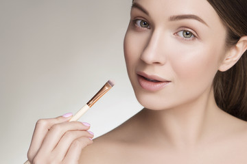 Obraz premium Beautiful brunette caucasian young woman prepare herself, applying lipstick on her lips with a brush. Clean, fresh, natural, flawless skin. Soft smile on her face. Close up on a neutral background