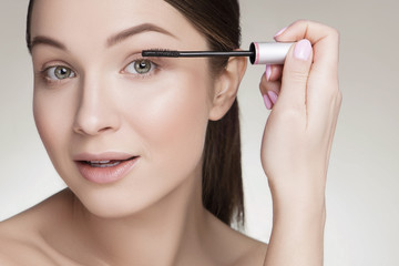 Fototapeta premium Beautiful brunette caucasian young woman prepare herself, applying mascara on her eyes with a brush. Clean, fresh, natural, flawless skin. Soft smile on her face. Close up on a neutral background