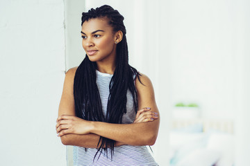 Portrait of pensive young African-American woman wearing striped tank top and jeans standing with folded arms leaning on wall - Powered by Adobe