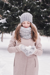 Female teenager holding heart shaped snow ball. Young girl in knitted scarf, mittens and hat standing in winter forest. Winter mood. Winter time