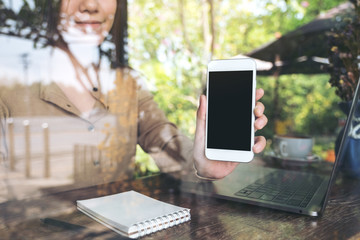 Mockup image of an Asian business woman holding and showing white mobile phone with blank black screen while using laptop in cafe