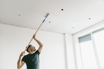 the man paints the ceiling in a gray color with a roller in a bright room. Repair and painting. - obrazy, fototapety, plakaty