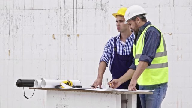 Two male engineers standing at table and using building blueprint to discuss project details when working at construction site