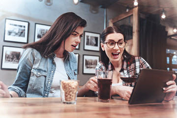 Look at this. Happy nice excited woman sitting with her friend and holding a tablet while showing her an interesting picture