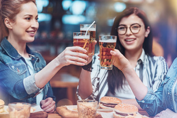 To us. Delighted pleasant pretty women holding glasses with beer and raising them while drinking at the pub