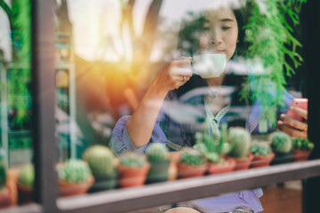 Asia beautiful woman using mobile phone and drinking coffee in the coffee shop on holiday.this photo take from outside the room pass the mirror.