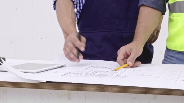 Close up shot of building blueprint lying on table while two male construction workers are discussing it and using pencils to add notes