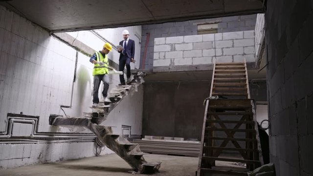 Two construction workers wearing hard hats going downstairs in unfinished building and discussing work progress