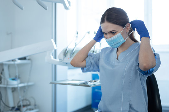 Taking safety measures. Charming female dentist putting on a face mask and wearing blue rubber gloves while getting ready for an appointment with a patient