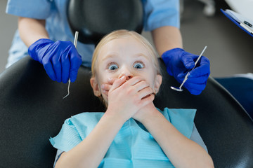 So scared. Pleasant little girl lying in a dentist chair and covering her mouth, being afraid of the checkup at the dentists