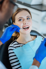 Caring about teeth. The close up of a gorgeous young woman smiling at her dentist and having her teeth examined by him with the help of a mouth mirror