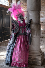 Fototapeta na wymiar Masked lady in ornate pink costume dress and mask standing next to pillar at Venice Carnival. Lady has feathered plumes, holds a decorated fan and is wearing black gloves