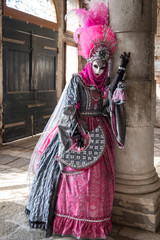 Fototapeta na wymiar Masked lady in ornate pink costume dress and mask standing next to pillar at Venice Carnival. Lady has feathered plumes, holds a decorated fan and is wearing black gloves