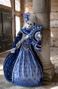 Masked woman holding ornate fan and wearing decorative blue dress standing next to an old stone column during Venice Carnival (Carnivale di Venezia)
