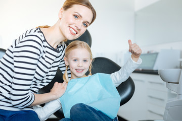 Fototapeta premium Perfect mood. Loving young mother hugging her little daughter and posing for the camera together with her while the girl sitting in the dentist chair and waiting for a checkup