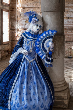 Masked woman holding ornate fan and wearing decorative blue dress standing next to an old stone column during Venice Carnival (Carnivale di Venezia)