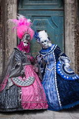 Fototapeta na wymiar Two women in masks and ornate blue and pink costumes standing in front of an old blue door at the end of an alley in Venice during the carnival (Carnivale di Venezia).